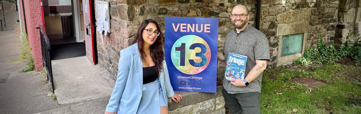Vanesa Kelly and Ian Garrett stand outside Harry Younger Hall with a Venue 13 sign and the 2025 Fringe guide.